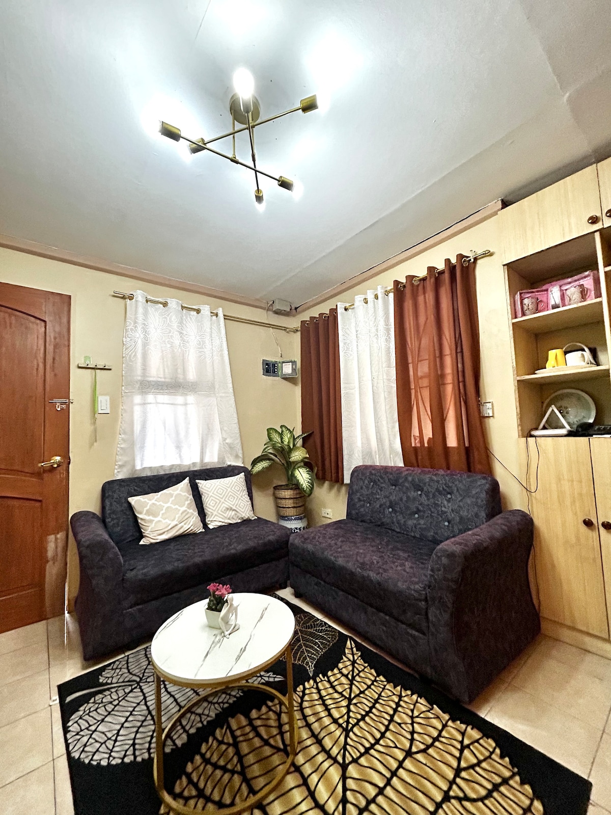 A cozy living room features two dark sofas complemented by a round coffee table with a leaf design. Natural light enters through a partly draped window, while decorative curtains add warmth. A small plant and shelving unit contribute to the stylish decor.