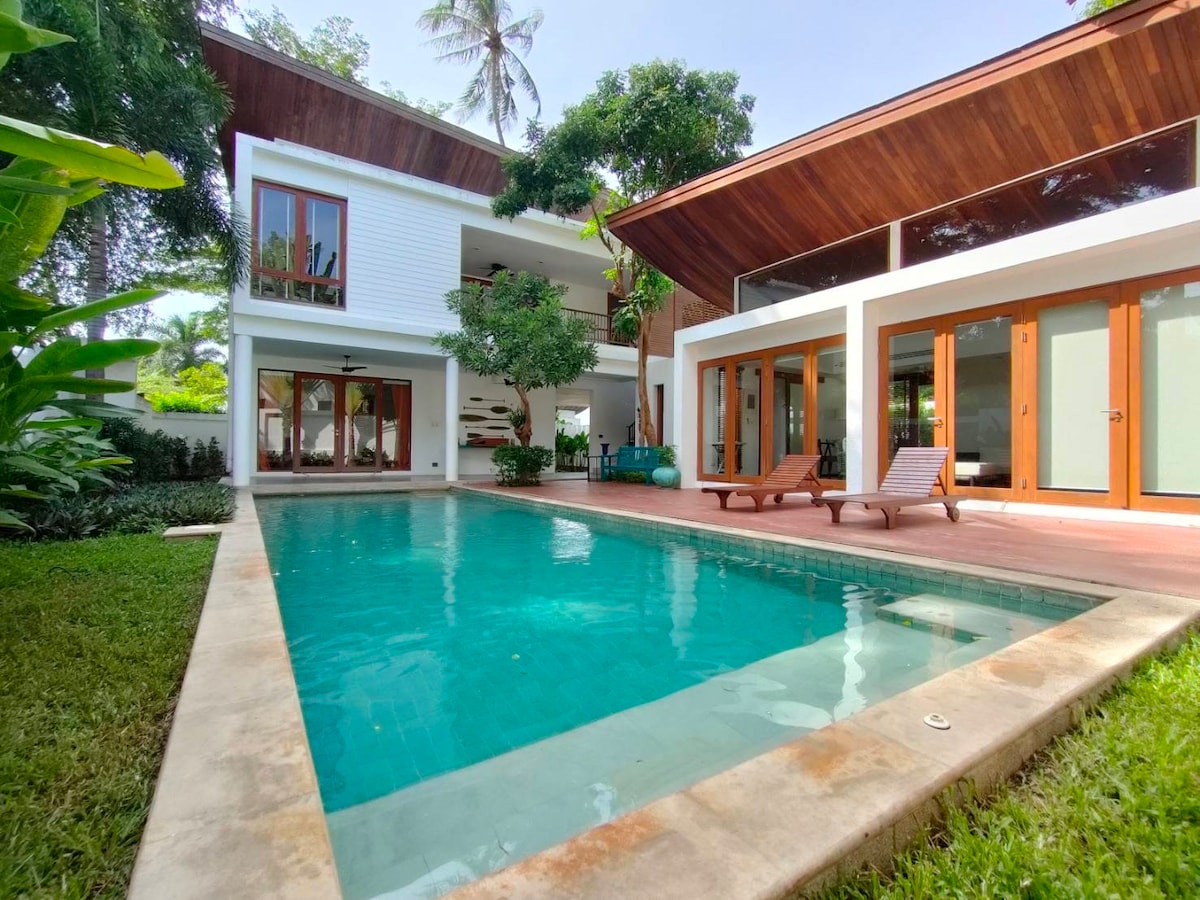 A private swimming pool is surrounded by a well-maintained garden, featuring lounge chairs positioned alongside. The two-story villa, characterized by large glass doors and a wooden roof, integrates indoor and outdoor living spaces harmoniously.