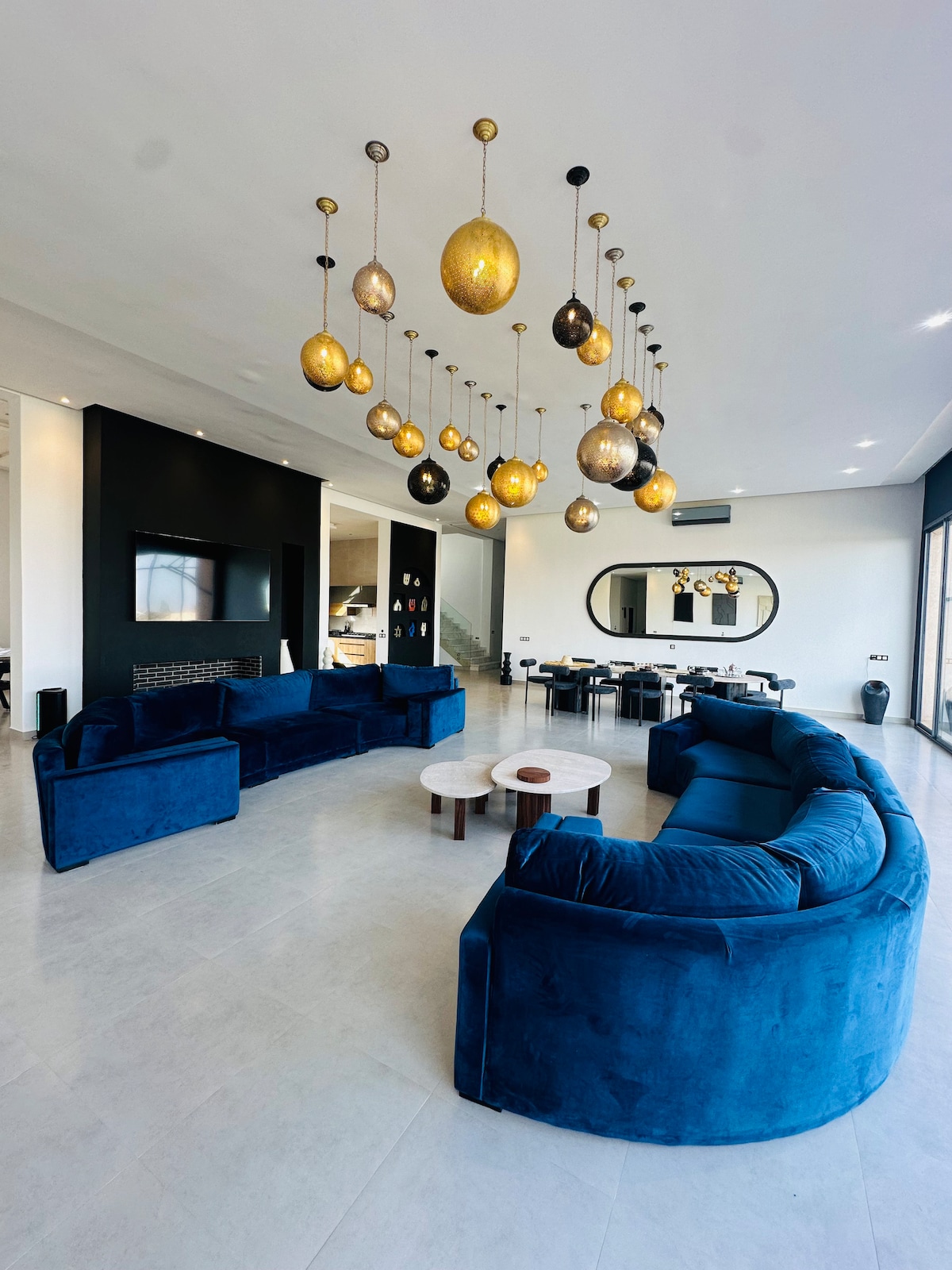 A spacious living area is furnished with modern blue sofas arranged in a circular layout, complemented by a centerpiece table. A decorative light fixture hangs from the ceiling, featuring varying sizes and colors. Large windows allow natural light to fill the room, enhancing the open atmosphere.