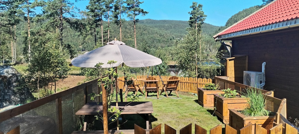 A serene outdoor space features wooden seating arrangements under a large umbrella. Raised garden beds are positioned around the grassy area, offering a peaceful setting surrounded by trees and mountainous views. A wooden fence encloses the yard, providing both privacy and a cozy atmosphere.