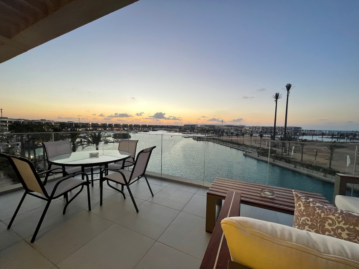 An outdoor terrace features a dining table with four chairs, accompanied by a leisure seating area. The view encompasses the marina and sea, while a colorful sunset casts a warm glow over the landscape, with palm trees lining the waterfront.