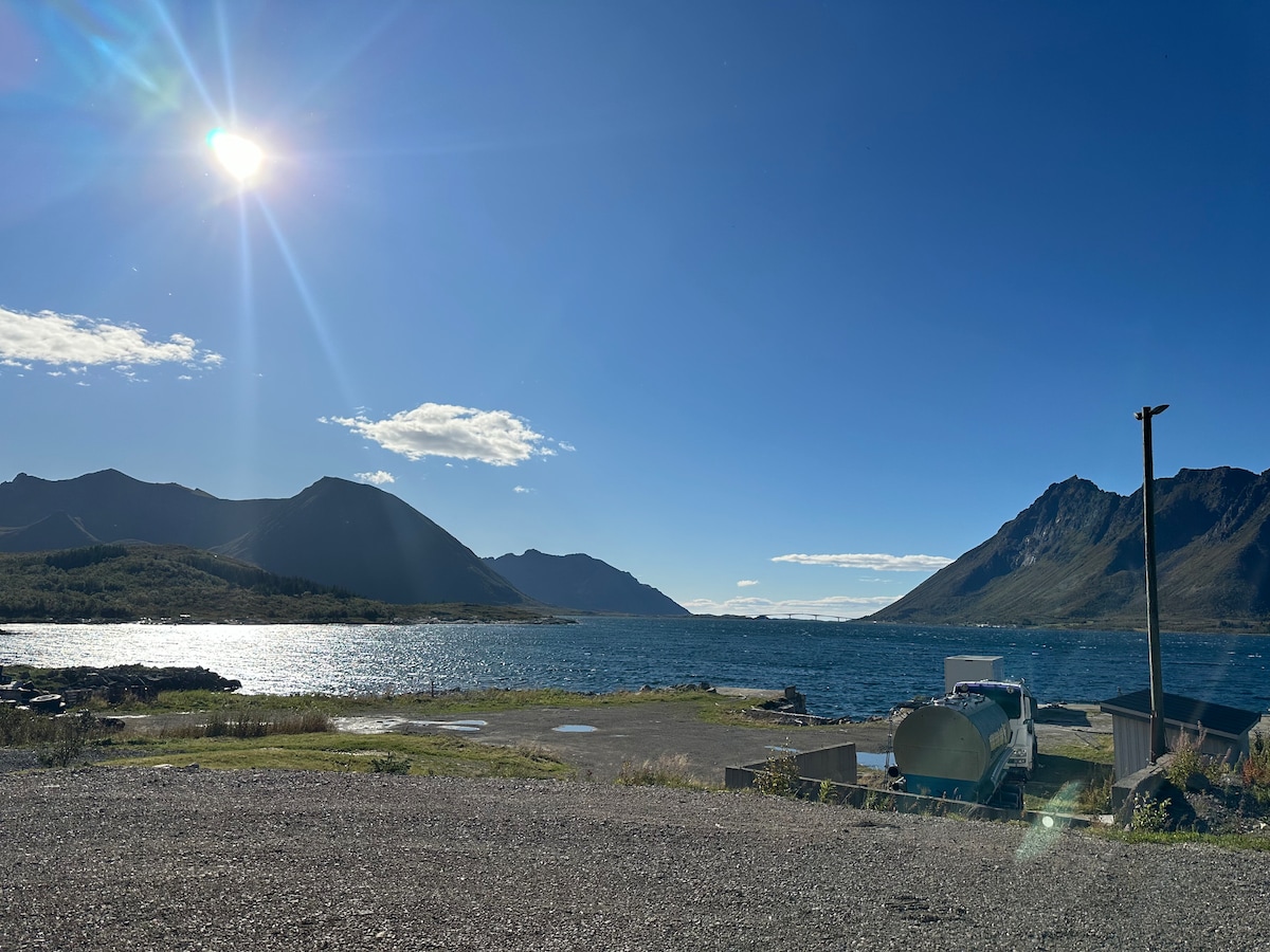A serene landscape is captured, showcasing a body of water reflecting sunlight under a clear blue sky. Surrounding mountains are visible in the distance, creating a tranquil backdrop. The scene is complemented by a gravel area with a vehicle parked nearby, suggesting accessibility.