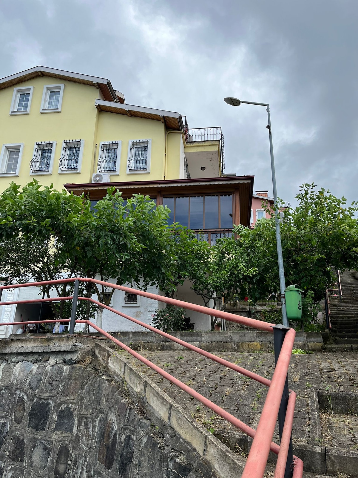 A multi-story building with yellow and white exteriors is set against a cloudy sky. A spacious balcony extends from the upper floor, while green trees line the pathway leading up to the entrance. A street lamp stands near the pathway's edge.