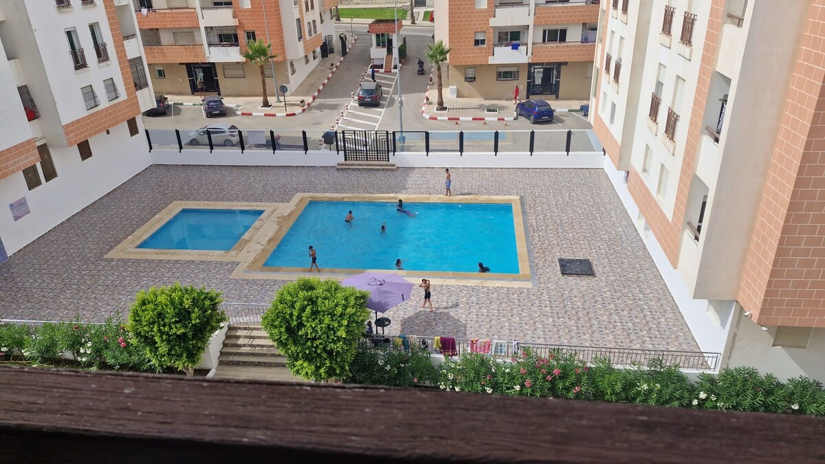 An outdoor pool area is visible, featuring a large rectangular pool and a smaller square pool. Several people can be seen enjoying the water. Surrounding the pool are tiled walkways and landscaped plants, while nearby buildings provide a view of the complex.