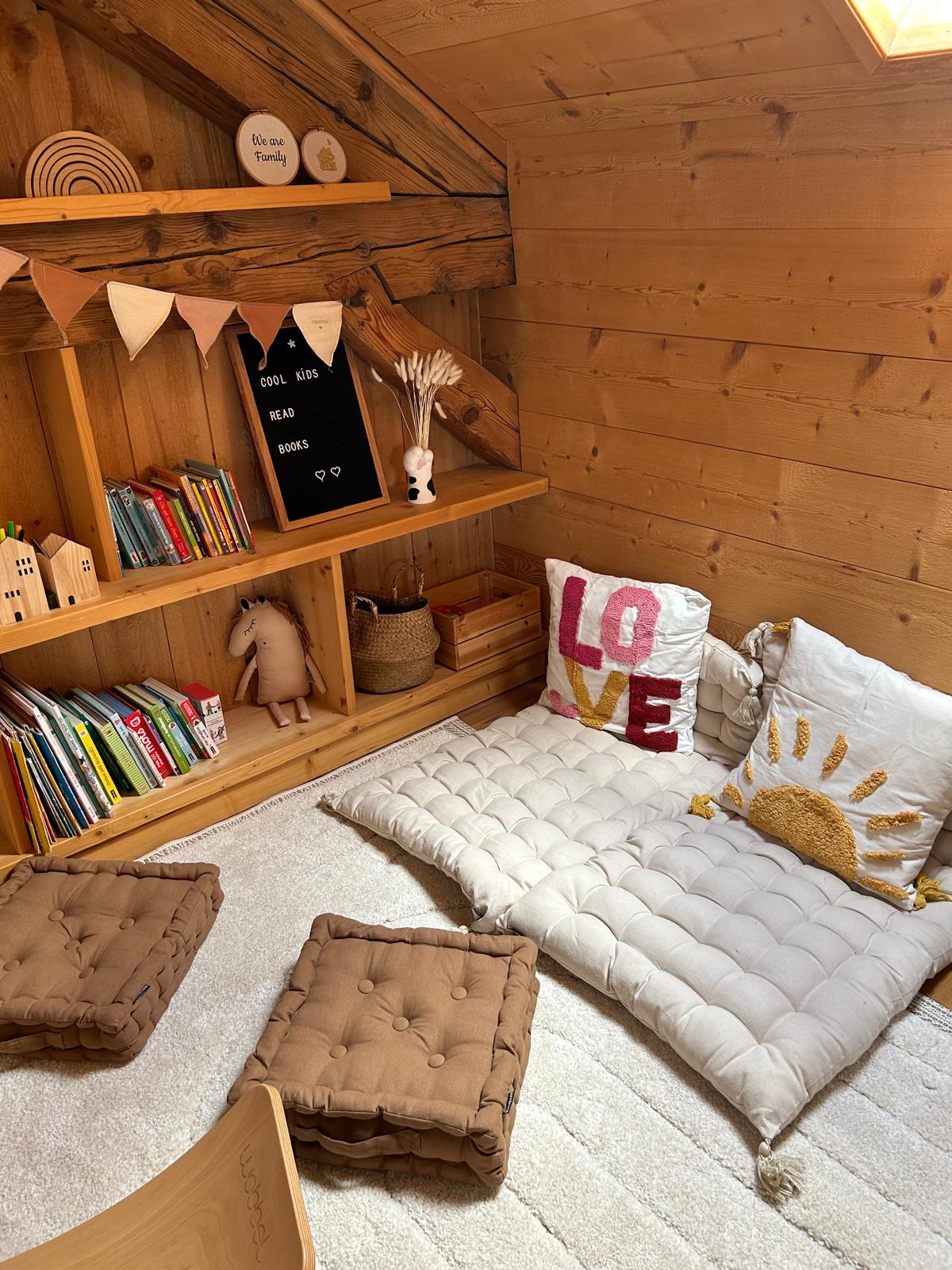 A play area designed for children is showcased, featuring cozy floor cushions and a plush rug. A wooden shelf is filled with books, and decorative items are displayed. Soft lighting enhances the warm wooden walls, creating a welcoming environment for imaginative play.