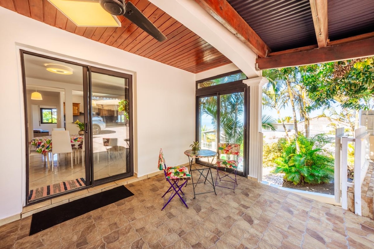 An outdoor seating area features two colorful chairs positioned beside a small table. Large glass doors open to the interior, with tropical greenery visible in the background, creating a relaxed and inviting entry space.
