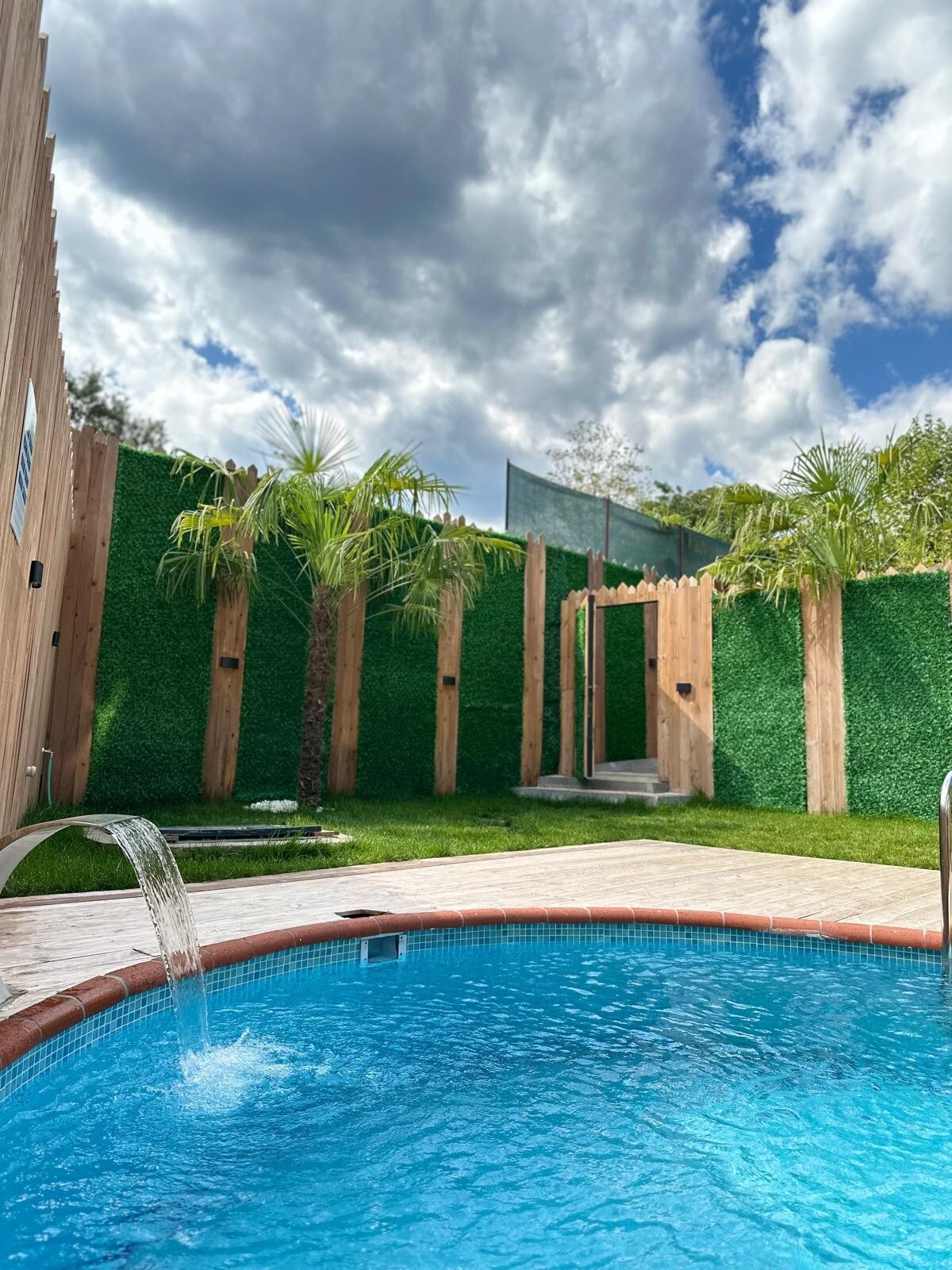 A heated pool is surrounded by lush greenery, featuring palm plants and wooden fencing. A gentle waterfall flows into the pool, creating a serene ambiance. The sky above is partly cloudy, with sunlight illuminating the outdoor space.