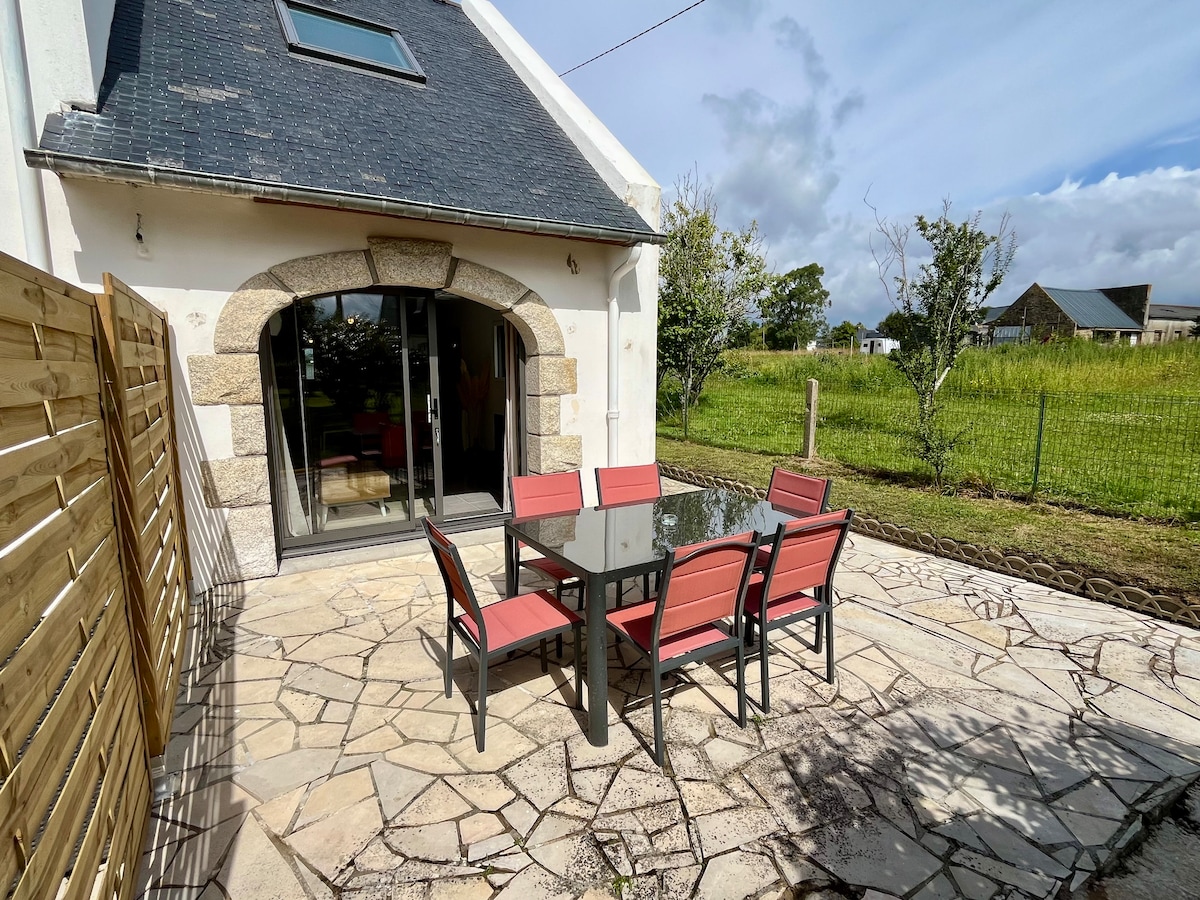 An outdoor terrace features a stone-paved floor and a dining set with six red chairs surrounding a glass table. The space is bordered by wooden fencing and open green fields, providing a calm setting for meals or relaxation.
