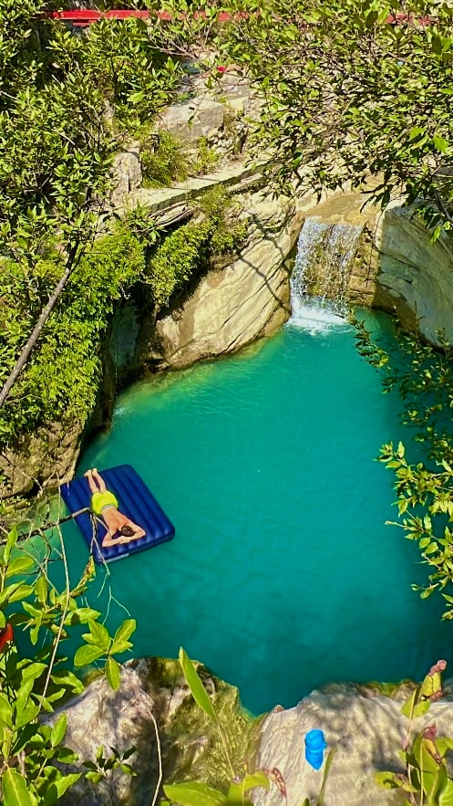 A natural pool is framed by rugged stone edges and lush greenery. Clear turquoise water reflects the sunlight, while a small waterfall cascades into the pool, creating a tranquil ambiance. A float is visible in the water, suggesting a relaxing experience in this serene setting.
