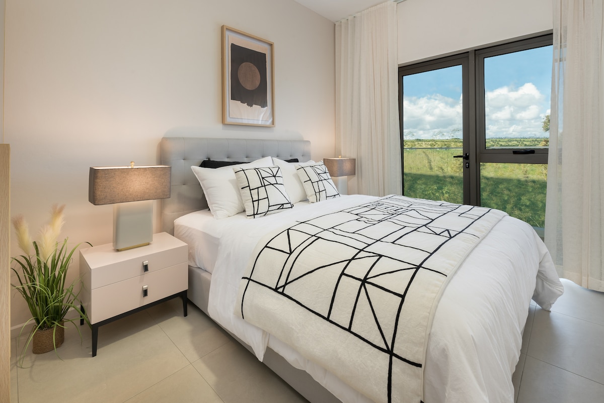 A restful bedroom features a spacious bed with a geometric patterned blanket. Two bedside tables with lamps sit on either side, and a window offers a view of the outdoor landscape. Soft curtains frame the window, adding a touch of warmth to the space.