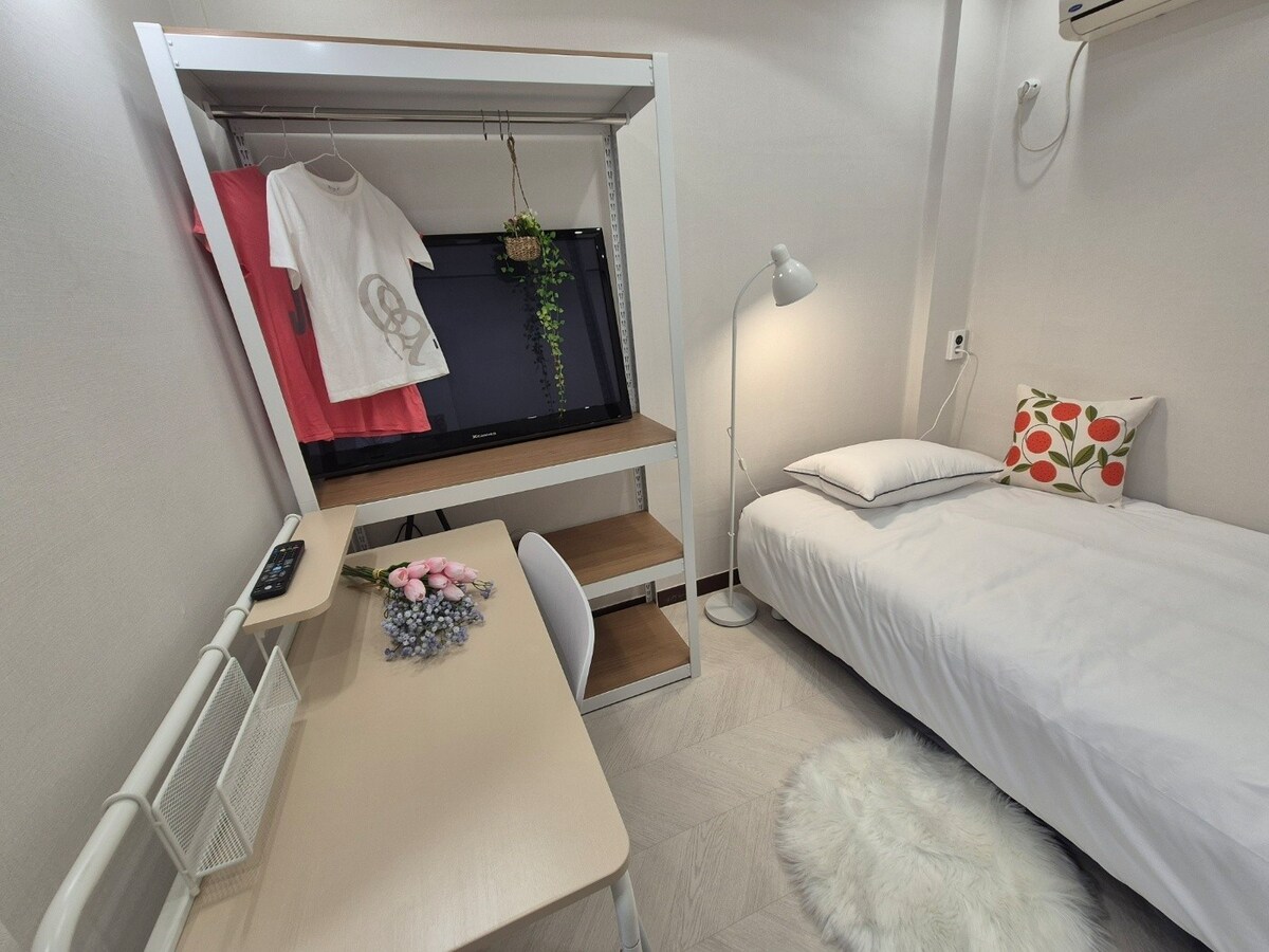 A compact sleeping area features a small bed with white bedding and a decorative pillow. A minimalist desk with a chair is present, accompanied by a wall-mounted TV and hanging clothing. A floor lamp and soft rug add to the cozy atmosphere.