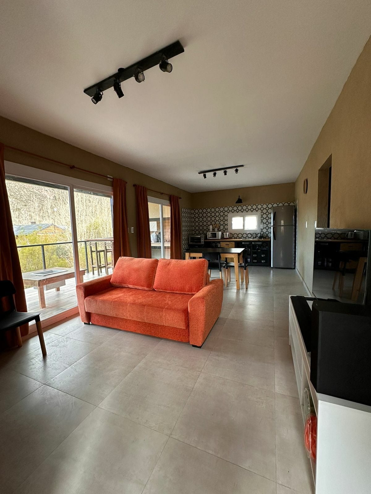 A spacious living area is furnished with an orange sofa and a black dining table set for four. Large glass sliding doors provide natural light and access to a terrace. A well-equipped kitchen is visible in the background, complemented by neutral-toned walls and modern lighting.