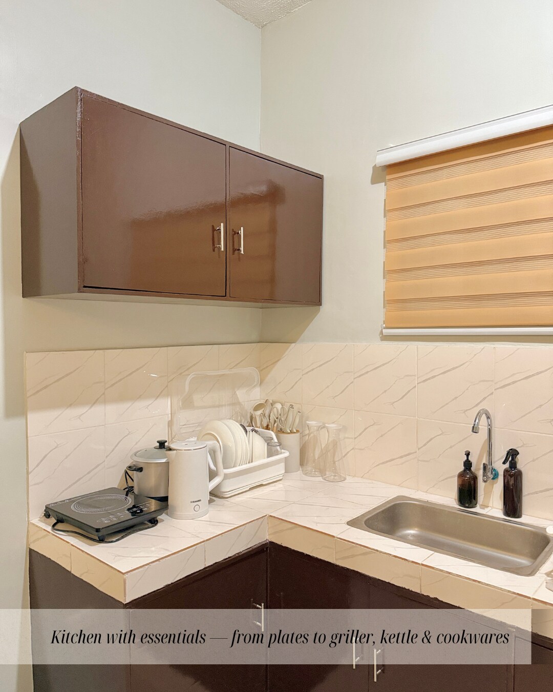 A kitchen space is equipped with essential items, including a stove, electric kettle, and various cookwares. The cabinets above store plates and glasses, while the sink area features black soap dispensers. Natural light enters through a window dressed with light-colored blinds.