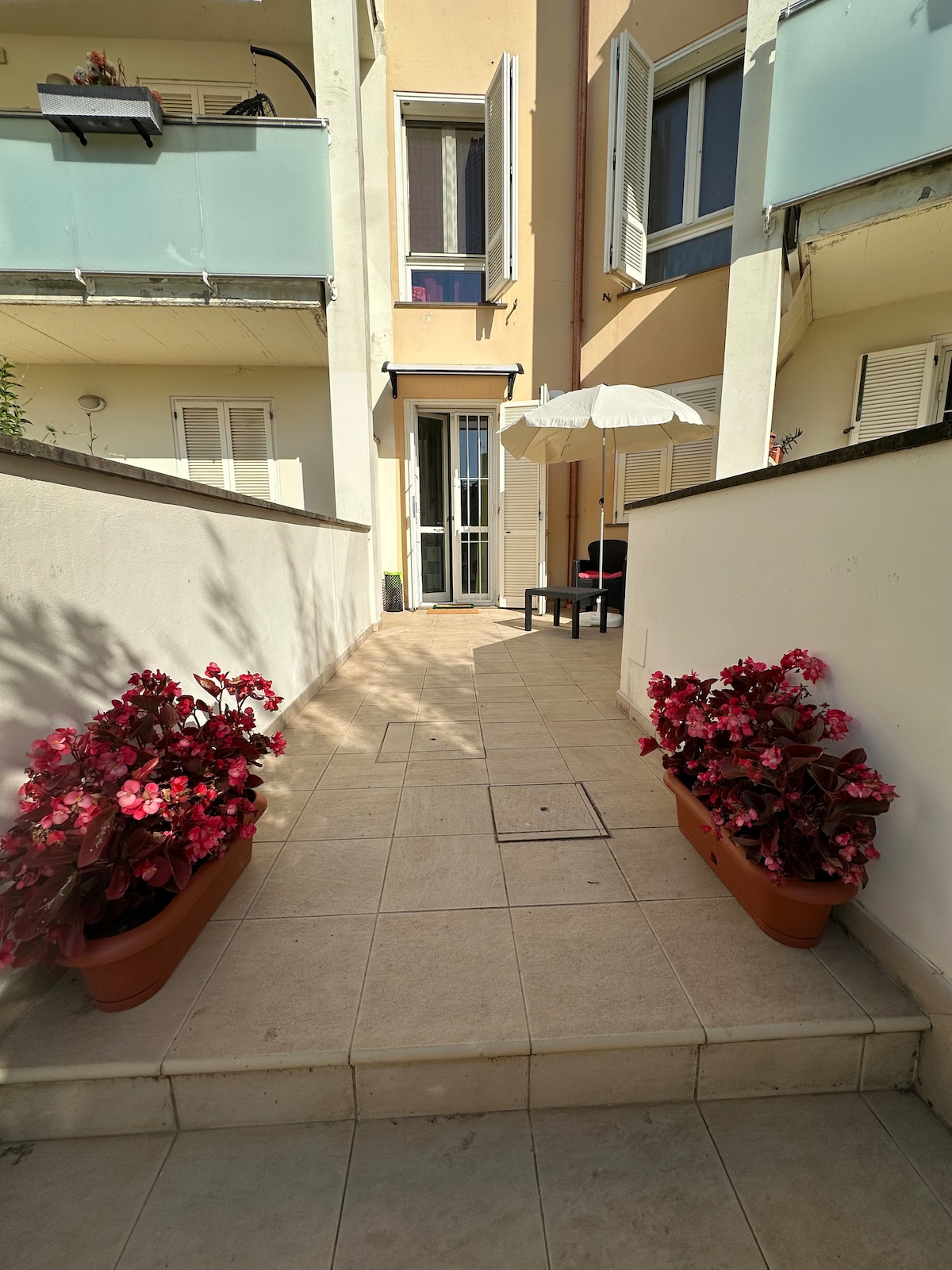 A pathway leads towards a private outdoor area, framed by flower pots with vibrant blooms. A small table and chairs are positioned under an umbrella, providing a shaded spot. Light tiles reflect soft sunlight, creating an inviting space for relaxation.