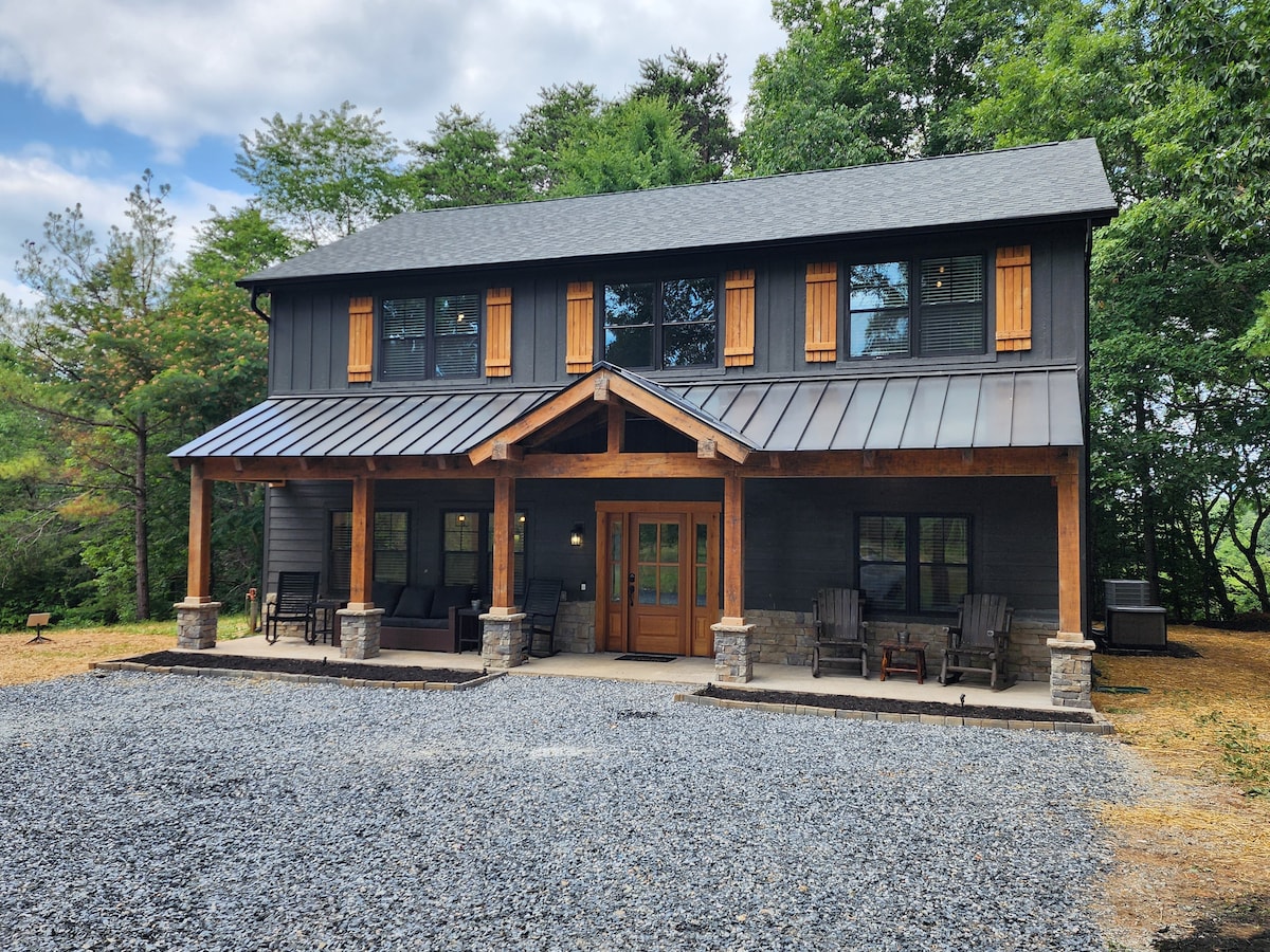 The exterior of a modern barndominium is showcased, featuring a dark wood facade with contrasting wooden shutters. A spacious porch is supported by wooden beams, with two seating areas visible. A gravel drive leads to the entrance, surrounded by trees and natural landscaping.