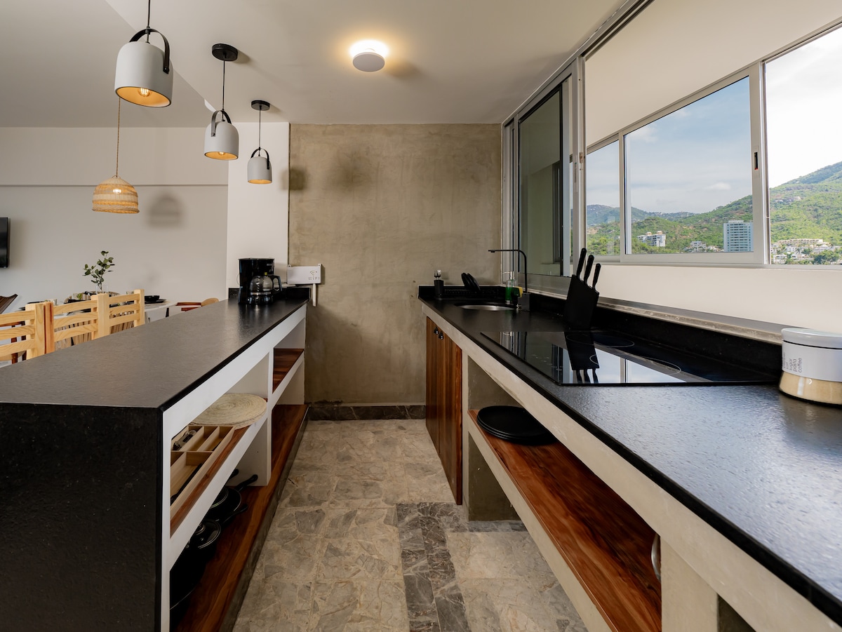 An open kitchen space features modern finishes with dark countertops and natural wood accents. Pendant lights illuminate the area above, while large windows offer a view of the outdoors. The kitchen is equipped with essential appliances and includes ample storage and preparation space.