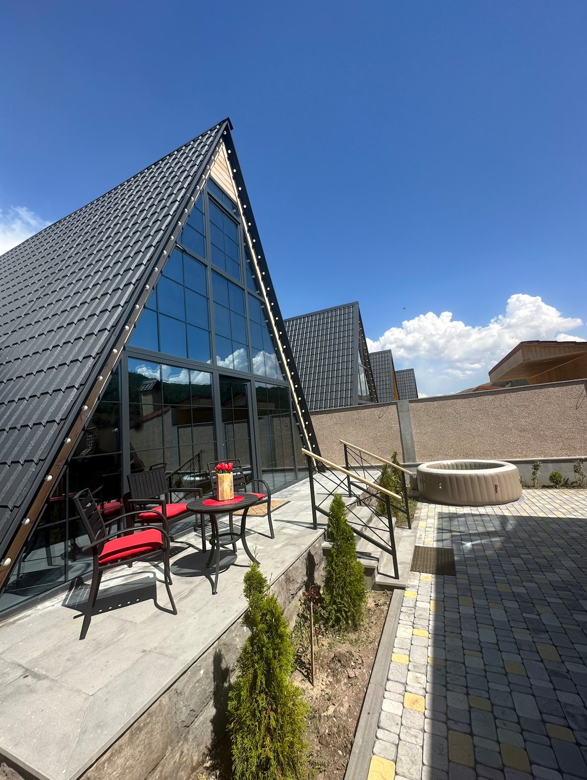 The modern structure features a distinctive triangular roof design with large windows reflecting natural light. An outdoor seating area is furnished with red chairs and a table. A hot tub is positioned nearby, surrounded by landscaped pathways and neat greenery.