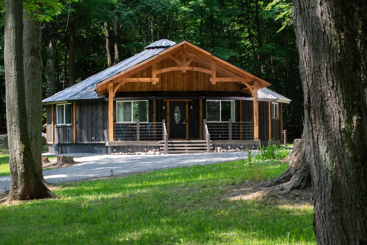 A modern wooden cabin is set among trees, featuring a large front porch with a symmetrical roof. The exterior showcases large windows, allowing natural light inside. A gravel driveway leads to the entryway, with a grassy area surrounding the cabin.
