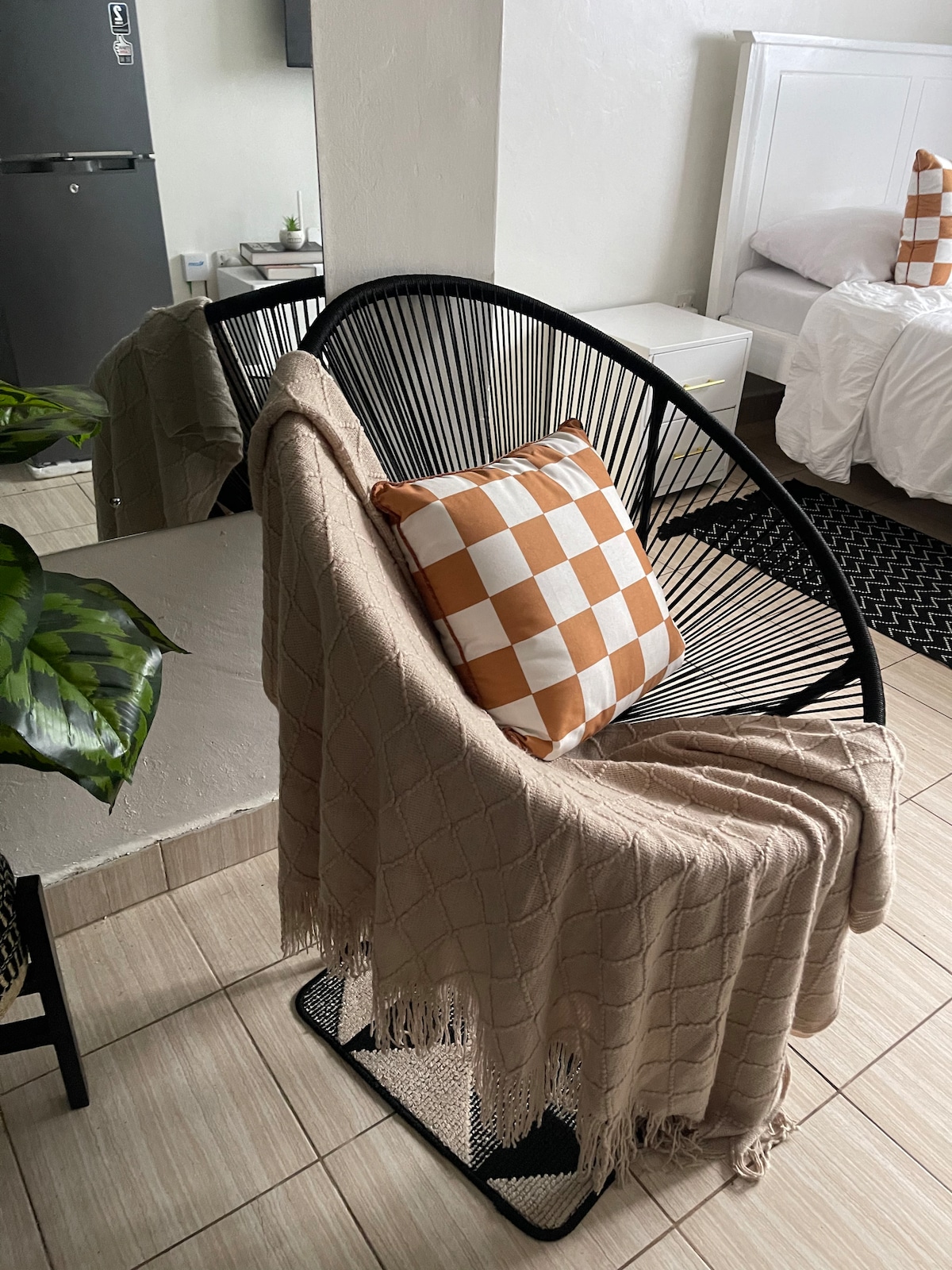 A black-accented lounge chair is positioned in a bright corner, adorned with a checkered cushion in warm tones. A soft, woven blanket drapes elegantly over the chair, enhancing the cozy appearance of the space, while light-colored tiles create a calming floor base.