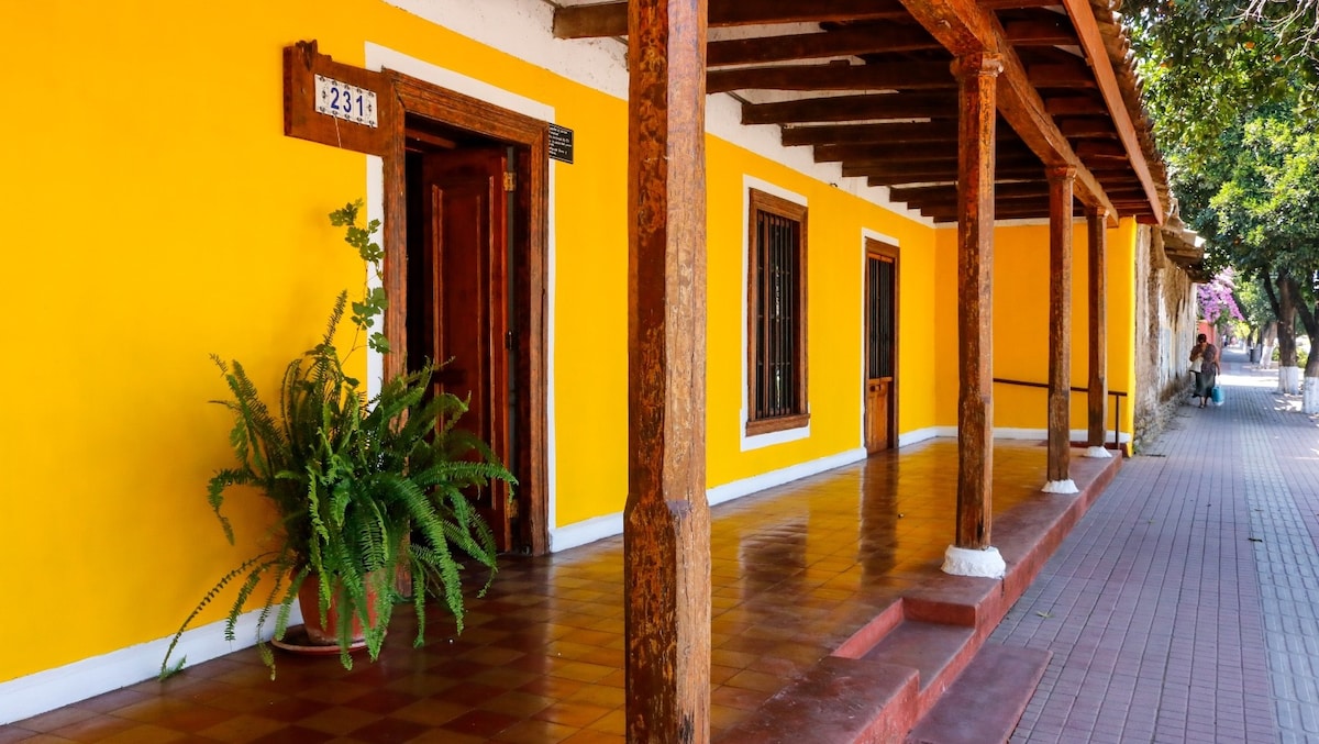 An exterior view of Hotel Los Artesanos showcases a vibrant yellow façade with traditional wooden beams. Potted plants add a touch of greenery to the entrance, while the tiled floor reflects the natural light. A quiet walkway lined with trees is visible in the background.