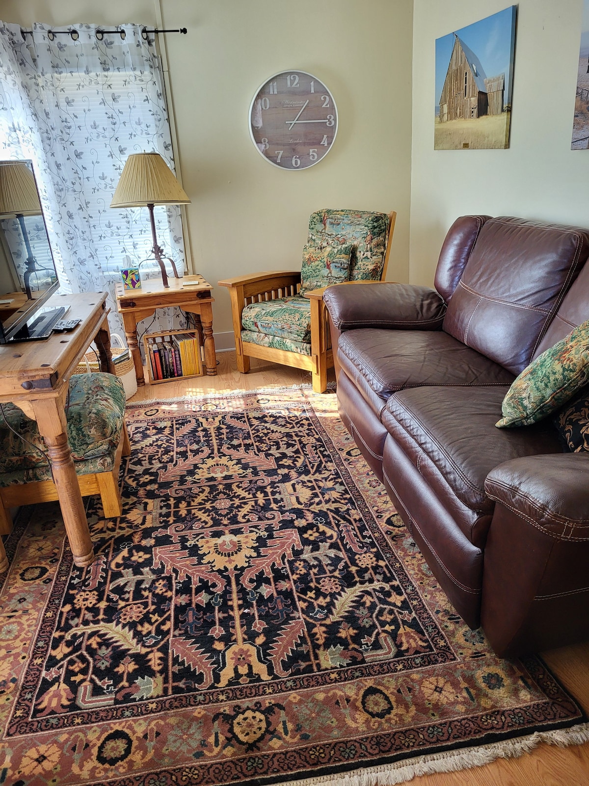 A cozy living area features a leather sofa paired with two armchairs, each adorned with floral upholstery. A wooden coffee table sits centrally on a decorative rug with intricate patterns. Natural light filters through sheer curtains, creating a warm ambiance.