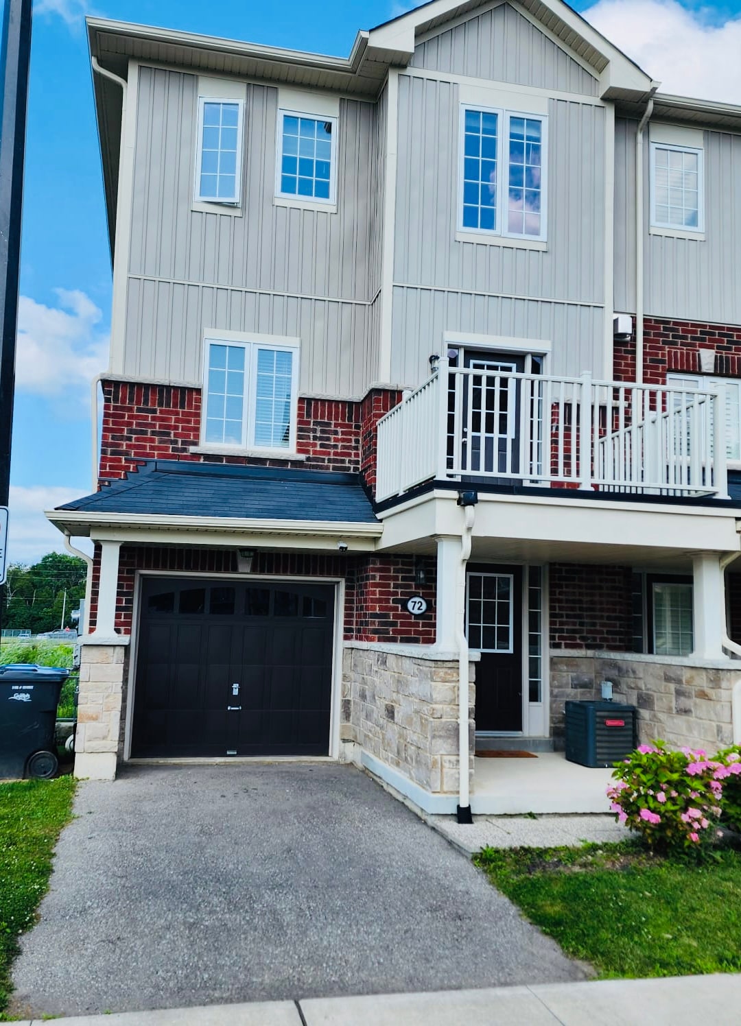 The exterior of the townhouse displays a combination of stone and brick materials, featuring a single-car garage with a dark door. A balcony is visible above the entrance, while vibrant flowers are present next to the porch, enhancing the welcoming appearance.