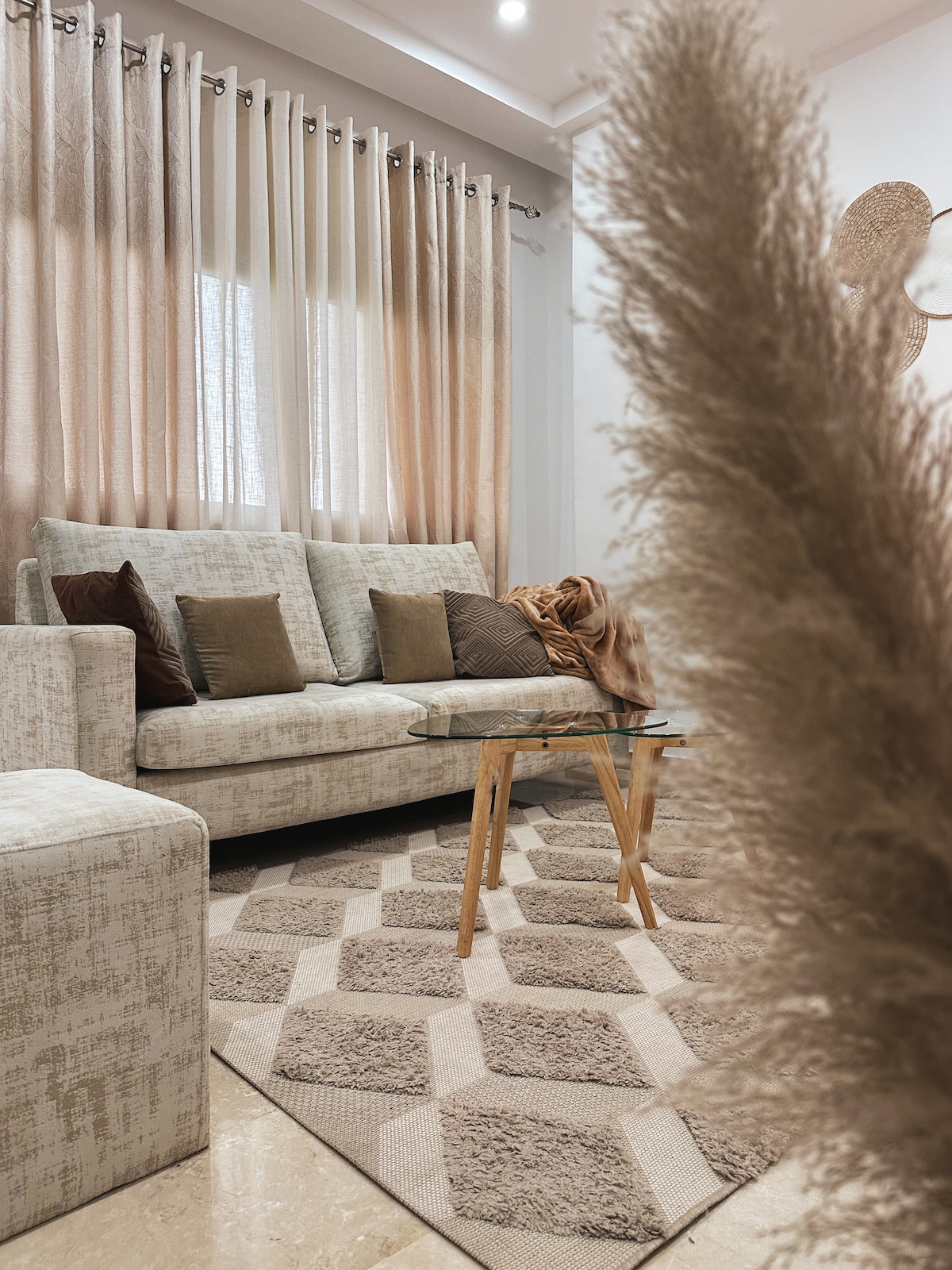 A light and airy living area features a comfortable sofa with decorative cushions and a glass coffee table. Soft textures are highlighted in the area rug below, while sheer curtains gently filter natural light from the windows.