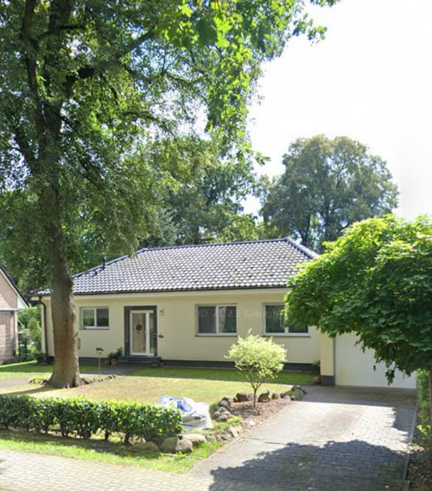 A single-family house is set in a serene environment surrounded by greenery. The front yard features neat hedges and a small tree, while a paved driveway leads to the entrance. Large windows allow natural light to enter the well-maintained structure.