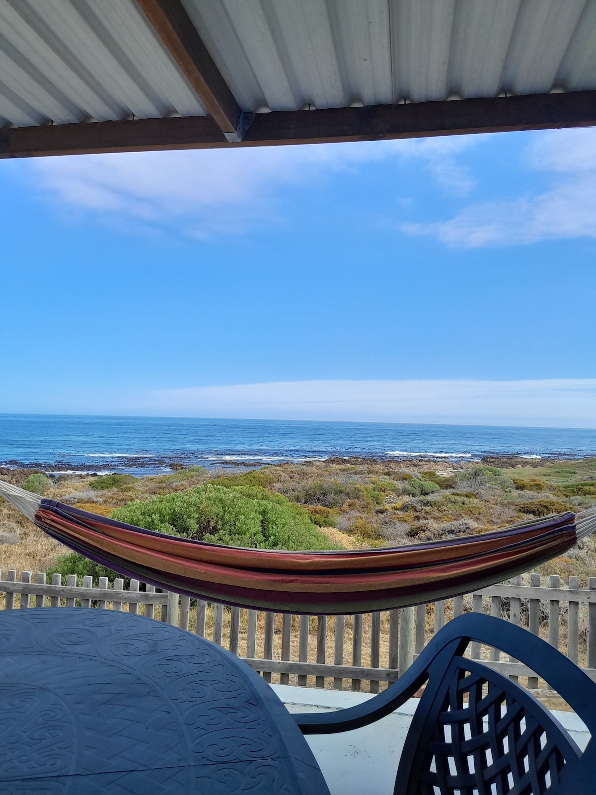 A hammock is suspended between two posts, offering a view of the ocean and coastline. In the foreground, a dark outdoor table and chair are visible, while the bright sky reflects the tranquil atmosphere of the coastal setting.