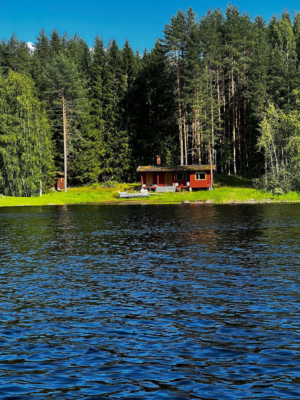 A red cabin is situated by the edge of a lake, surrounded by tall green trees. The cabin features a terrace with outdoor seating, providing a serene view of the water. Reflections of the foliage are visible on the calm surface of the lake.