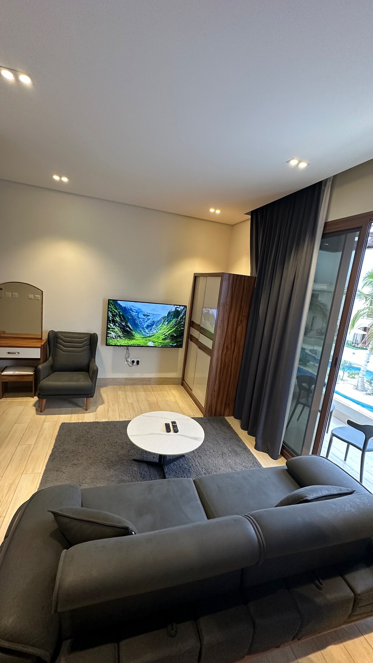 A modern living area is highlighted by a comfortable sectional sofa and a round coffee table. A large television is mounted on the wall, while large windows allow natural light to fill the space. Dark curtains provide options for light control.