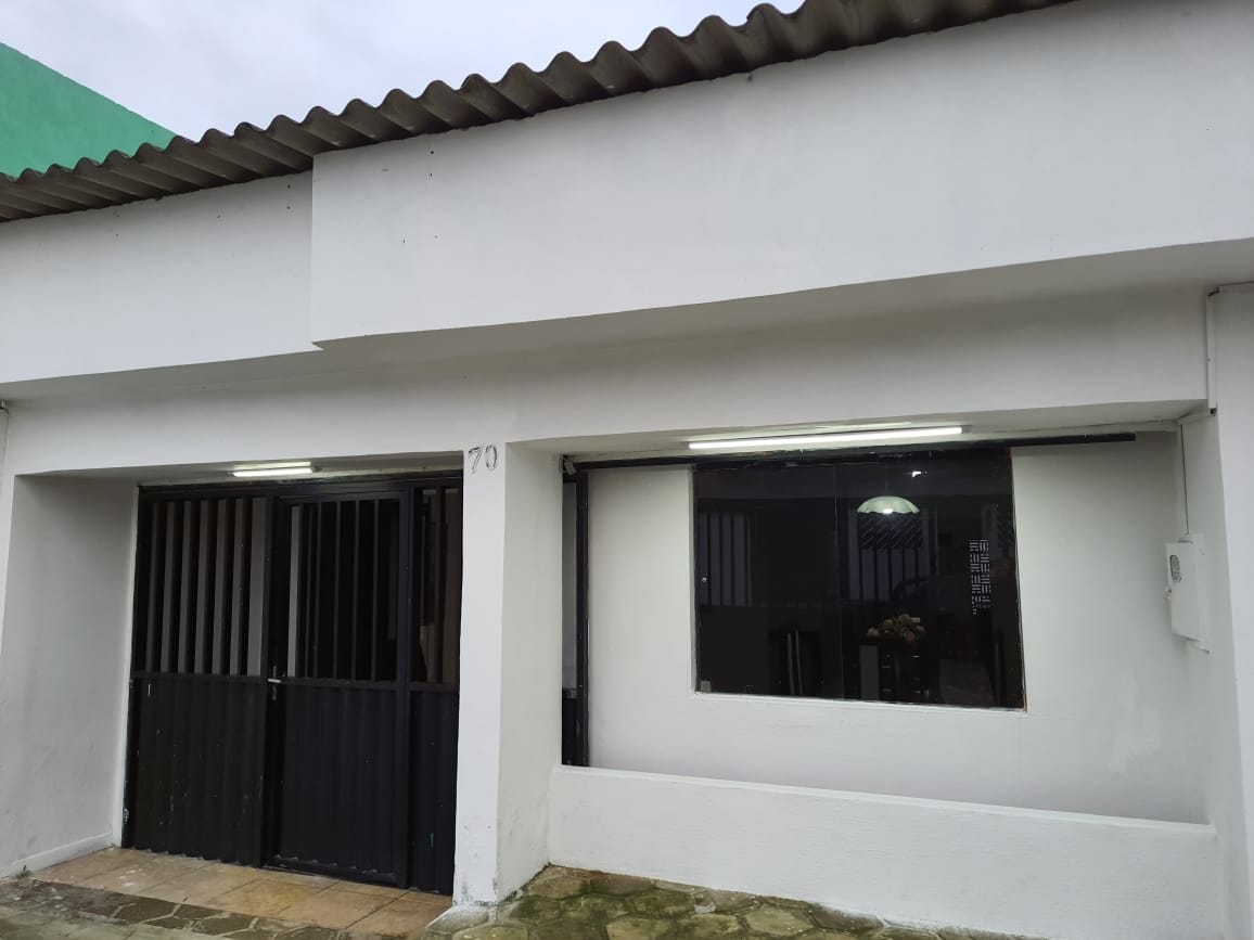 The exterior of the house features a simple façade with a white stucco finish. A black gate provides access, alongside large windows that allow natural light to enter. The tiled walkway leads to a smooth stone surface, contributing to a clean and accessible entrance.