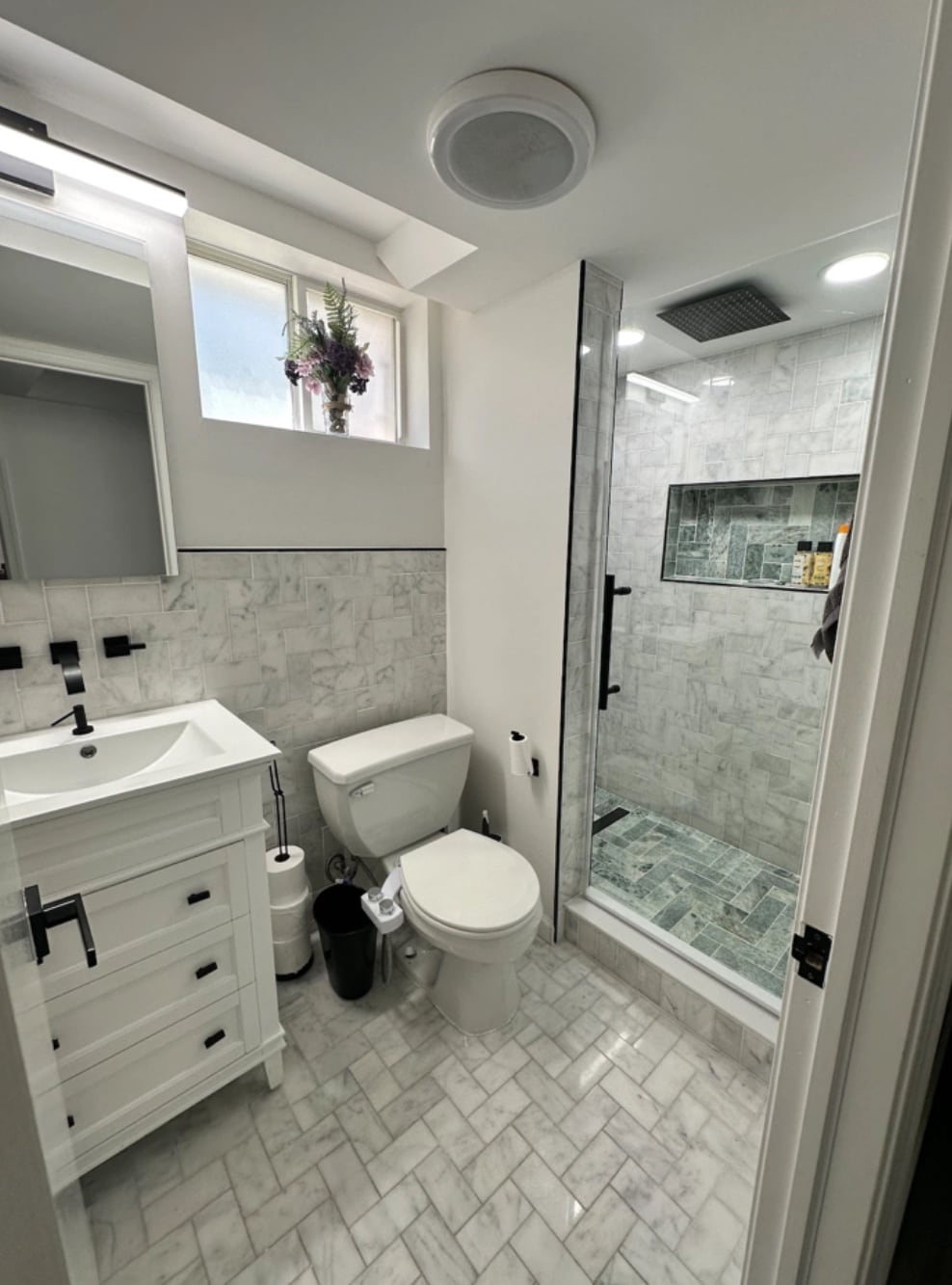 A clean bathroom features a separate glass shower with tiling. A white vanity is positioned beside a modern toilet. Natural light filters through a small window, adorned with plants. Marble tiles cover the floor, contributing to a bright and fresh ambiance.
