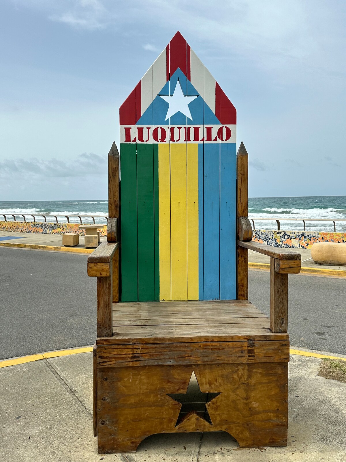 A colorful, oversized wooden throne is displayed, featuring a painted design in red, blue, and yellow with a star at the top. The throne is situated along a coastal walkway with waves visible in the background, providing a vibrant backdrop to the scene.