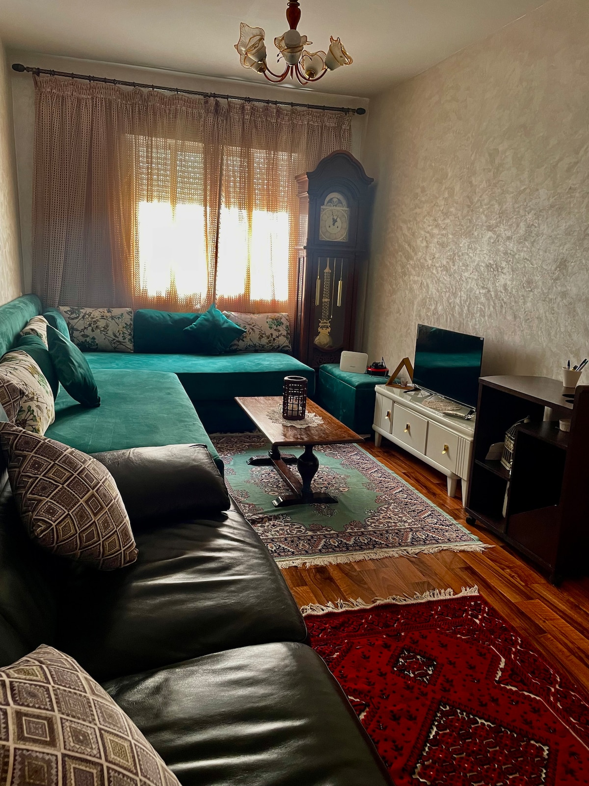 A cozy living area features a dark leather sofa, complemented by a teal sectional and patterned cushions. A vintage clock stands against the wall, while a small coffee table occupies the center. A television is set on a white console, with vibrant rugs adorning the wooden floor.