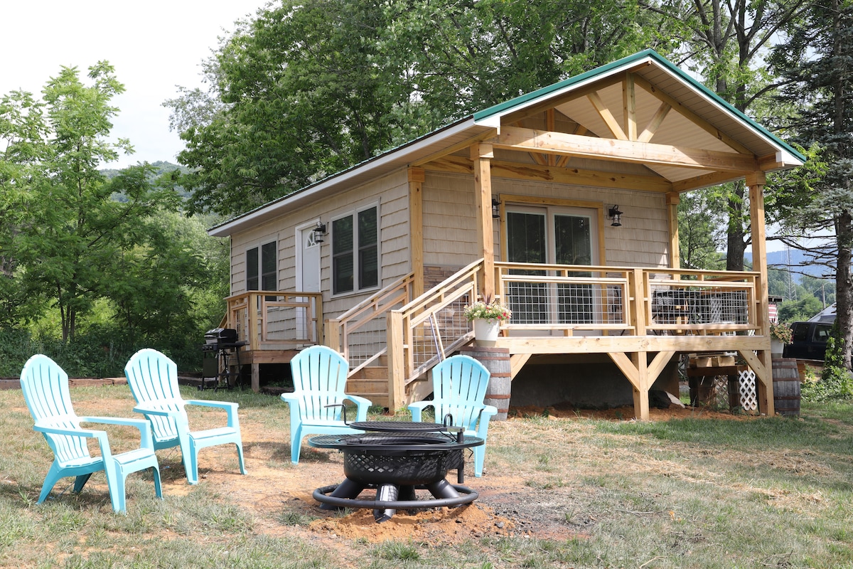 A cozy cabin exterior is highlighted by a welcoming front porch. Several blue Adirondack chairs surround a fire pit on grassy ground, creating an inviting outdoor space. Lush trees provide a natural backdrop, and large windows allow for ample natural light.