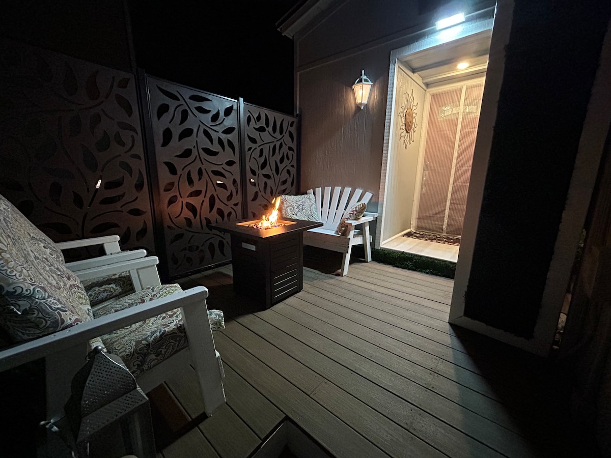 An outdoor deck area is highlighted by a propane fire pit, creating a warm focal point. Two chairs with patterned cushions are positioned nearby. A softly illuminated entrance can be seen in the background, adding a welcoming touch to the space.