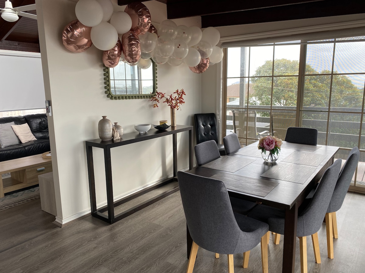 A dining area is featured with a rectangular wooden table surrounded by six gray upholstered chairs. A vase with flowers is placed at the center of the table. A mirror hangs on the wall, reflecting natural light from large windows that overlook a balcony.