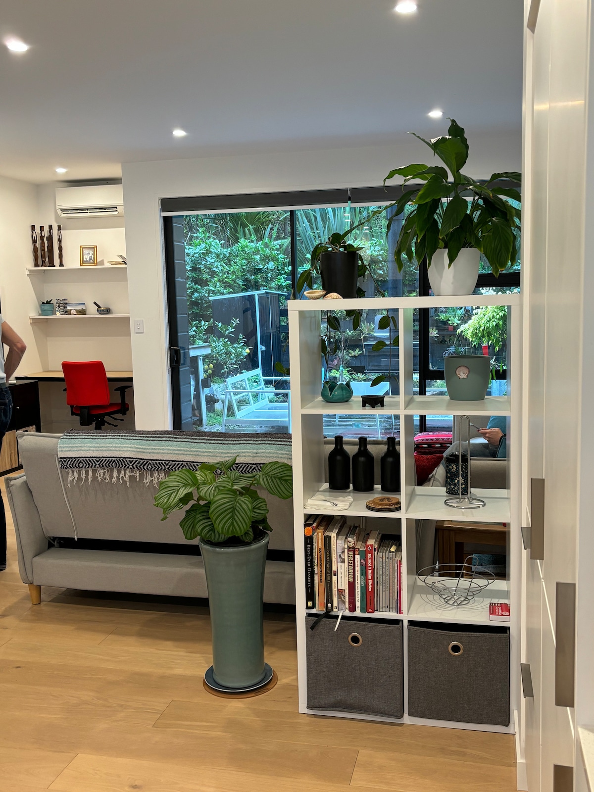 A modern living space features a gray sofa positioned beside a shelf displaying plants and decorative items. A view of a lush garden is visible through glass doors, enhancing the indoor-outdoor flow. Neutral colors create a relaxed atmosphere, complemented by wooden flooring.