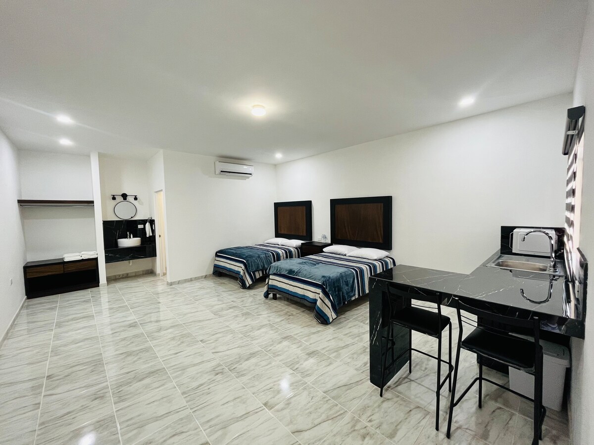 A spacious room features two large beds with dark wooden headboards, complemented by crisp bedding. A kitchenette area with a black countertop and two stools is visible. Light-colored tiled flooring and bright overhead lighting create a clean and open ambiance.