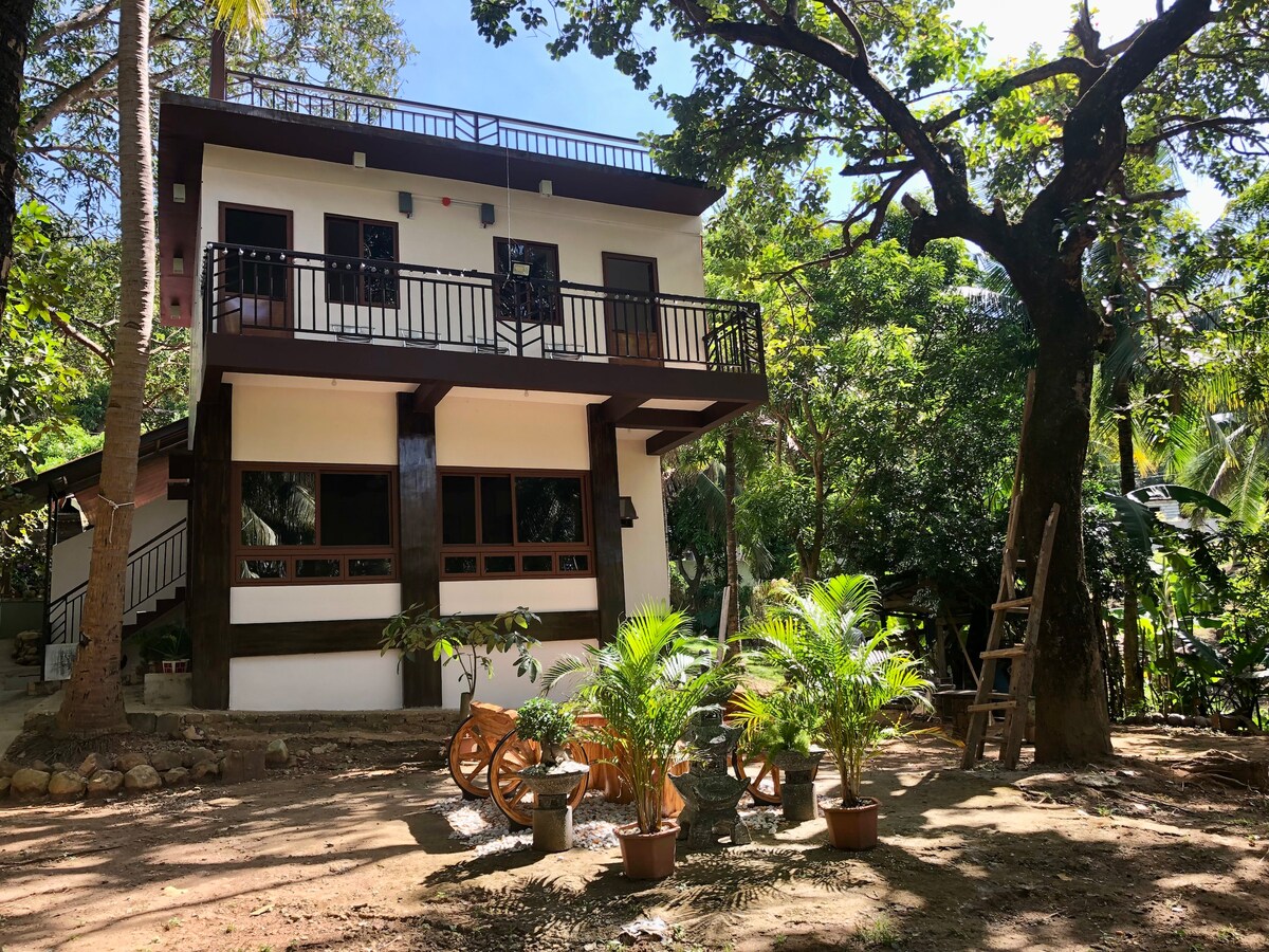 A two-story farmhouse is situated amidst lush greenery, featuring a wooden balcony on the upper level and large windows on the lower level. Potted plants and decorative elements are visible in the yard, creating a serene outdoor setting.