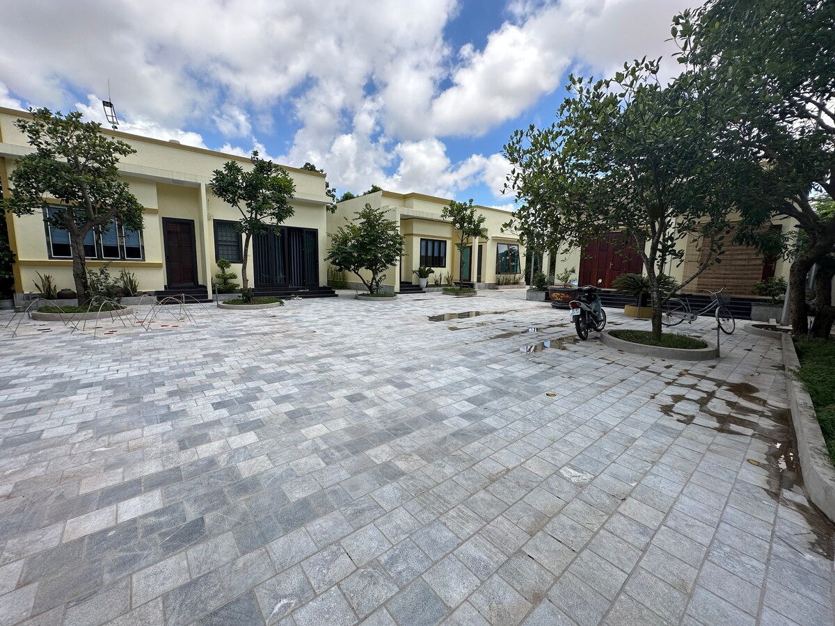 A spacious courtyard is paved with light grey tiles, lined by low-maintenance shrubs and small trees. Several individual units are visible, each with large windows and wooden doors. The area is bathed in natural light under a partly cloudy sky.