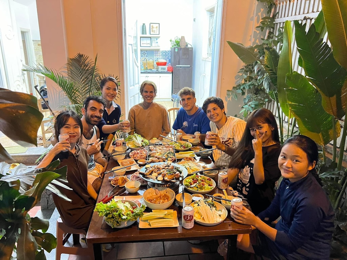 A spacious dining area is filled with a large wooden table set for a meal, surrounded by lush green plants. Various dishes of food, including vegetables and meats, are beautifully arranged. Guests are gathered around, enjoying the food and drinks in a warm, communal atmosphere.