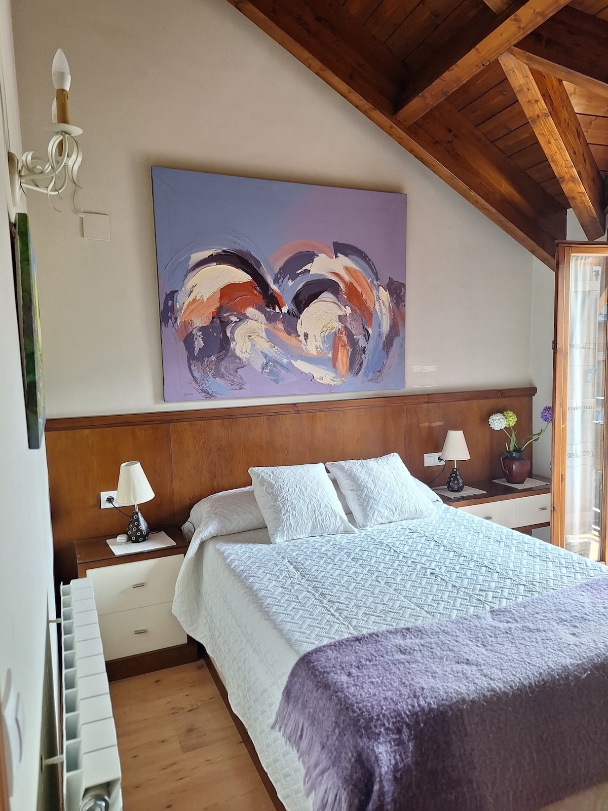 A cozy bedroom is presented with a light-colored quilt covering a spacious bed. Two bedside tables with lamps are positioned on either side. A colorful abstract artwork adorns the wall above the bed. Wooden beams and a large window add to the inviting ambiance.