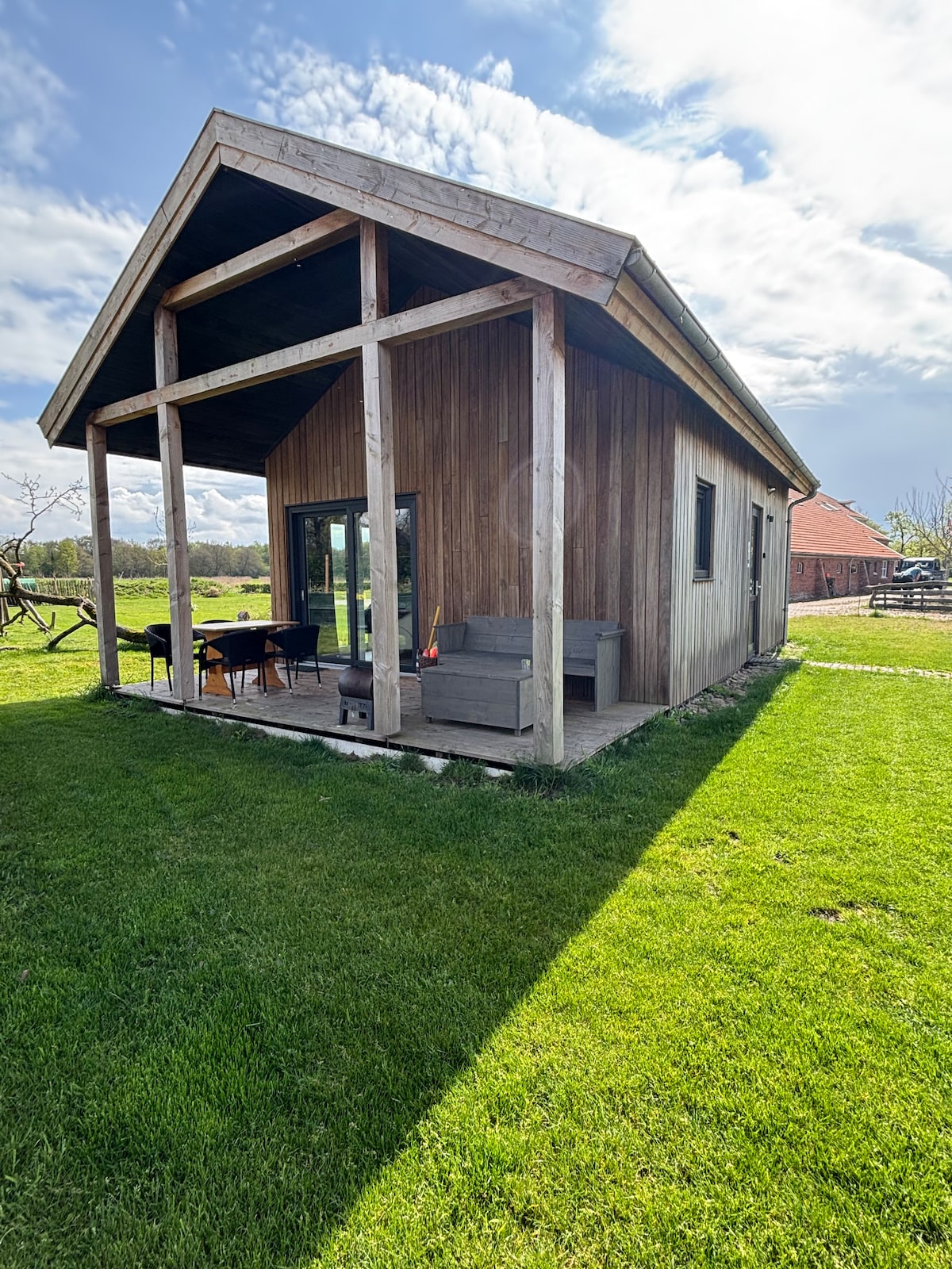 A private holiday home is viewed from the exterior, featuring a wooden façade and a spacious porch area. Outdoor seating is arranged on the porch, surrounded by green grass. The setting includes a backdrop of trees and the horizon, creating a sense of openness.