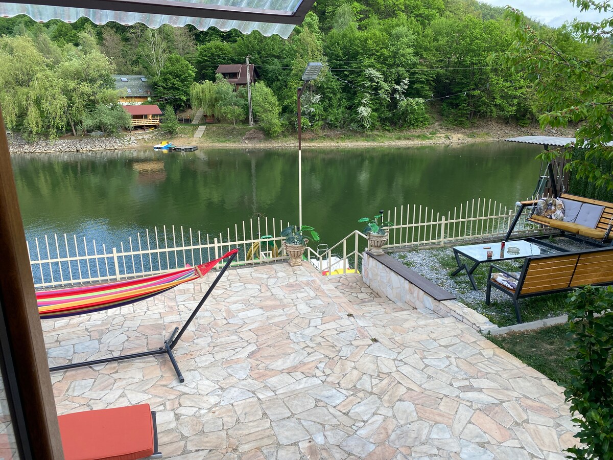 A spacious outdoor terrace is featured, overlooking a serene lake bordered by lush greenery. A hammock hangs between two posts, and a seating area with a table is visible beside the water. The area is surrounded by a wooden fence and includes gentle steps leading down to the lake.