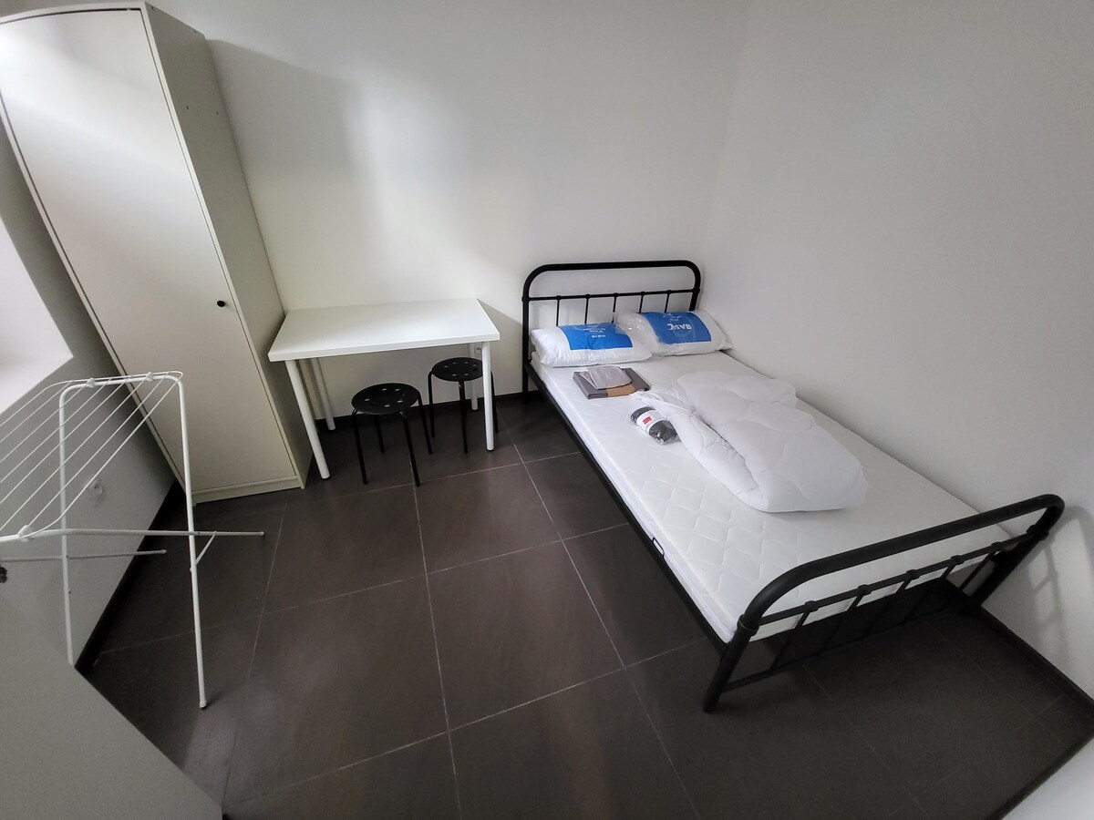 A simple sleeping area features a single metal frame bed with a mattress topped with a white comforter. A desk with two black chairs is positioned nearby, along with a freestanding clothing rack and a white storage cabinet situated against the wall.
