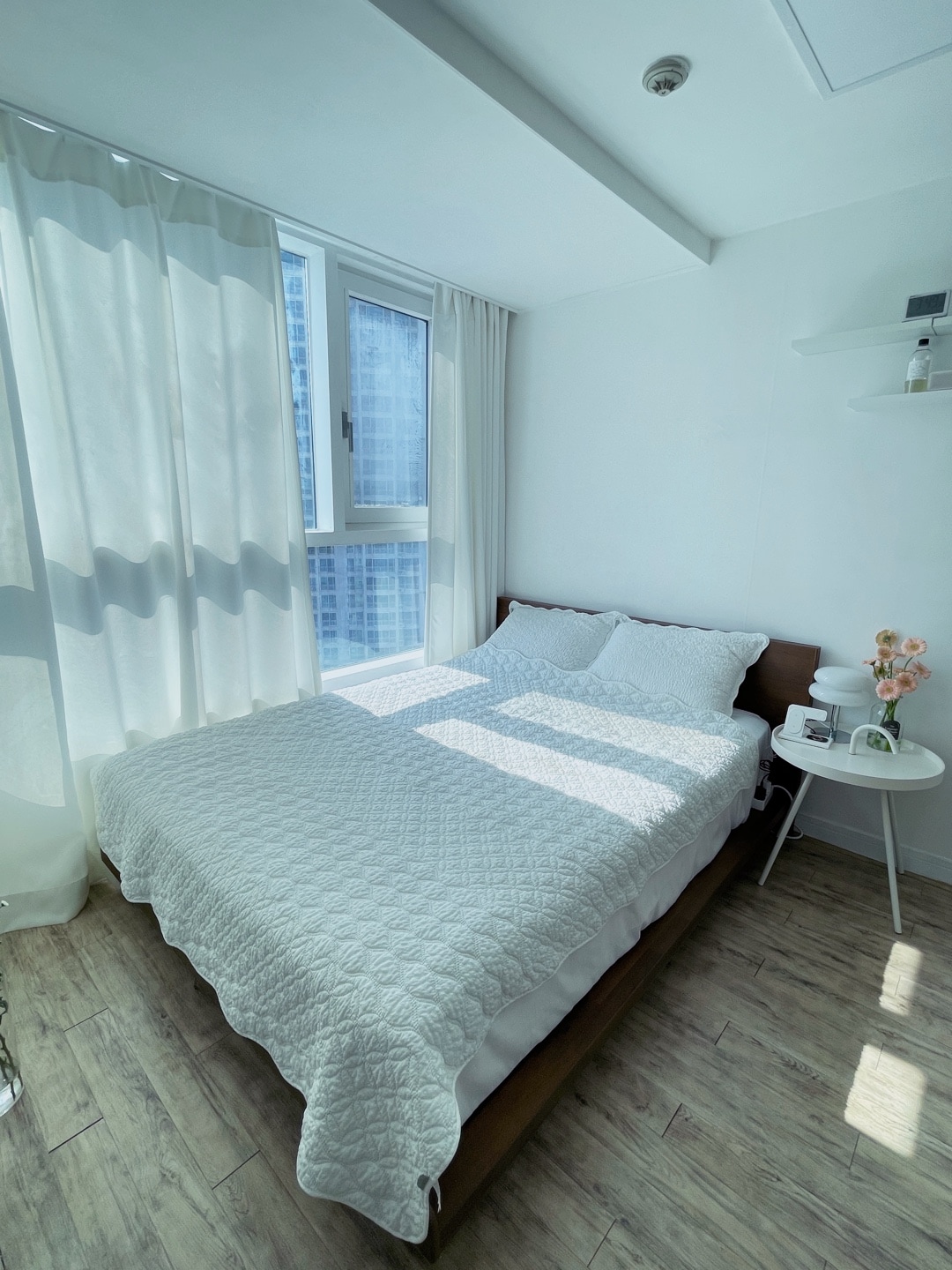 A bright bedroom features a spacious bed with a light quilt and crisp white linens, positioned beside a gently illuminated window. Soft curtains allow for filtered sunlight, creating a serene atmosphere. A small side table with a floral arrangement adds a touch of elegance.