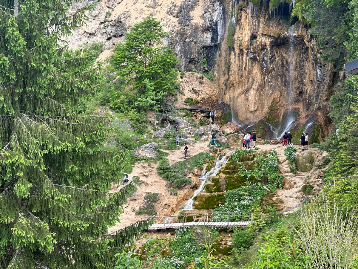 A natural landscape depicts a rocky terrain with cascading water flows from a cliff. Dense greenery surrounds the area, while visitors can be seen exploring the site. A pathway connects different levels of the terrain, leading through patches of vegetation.