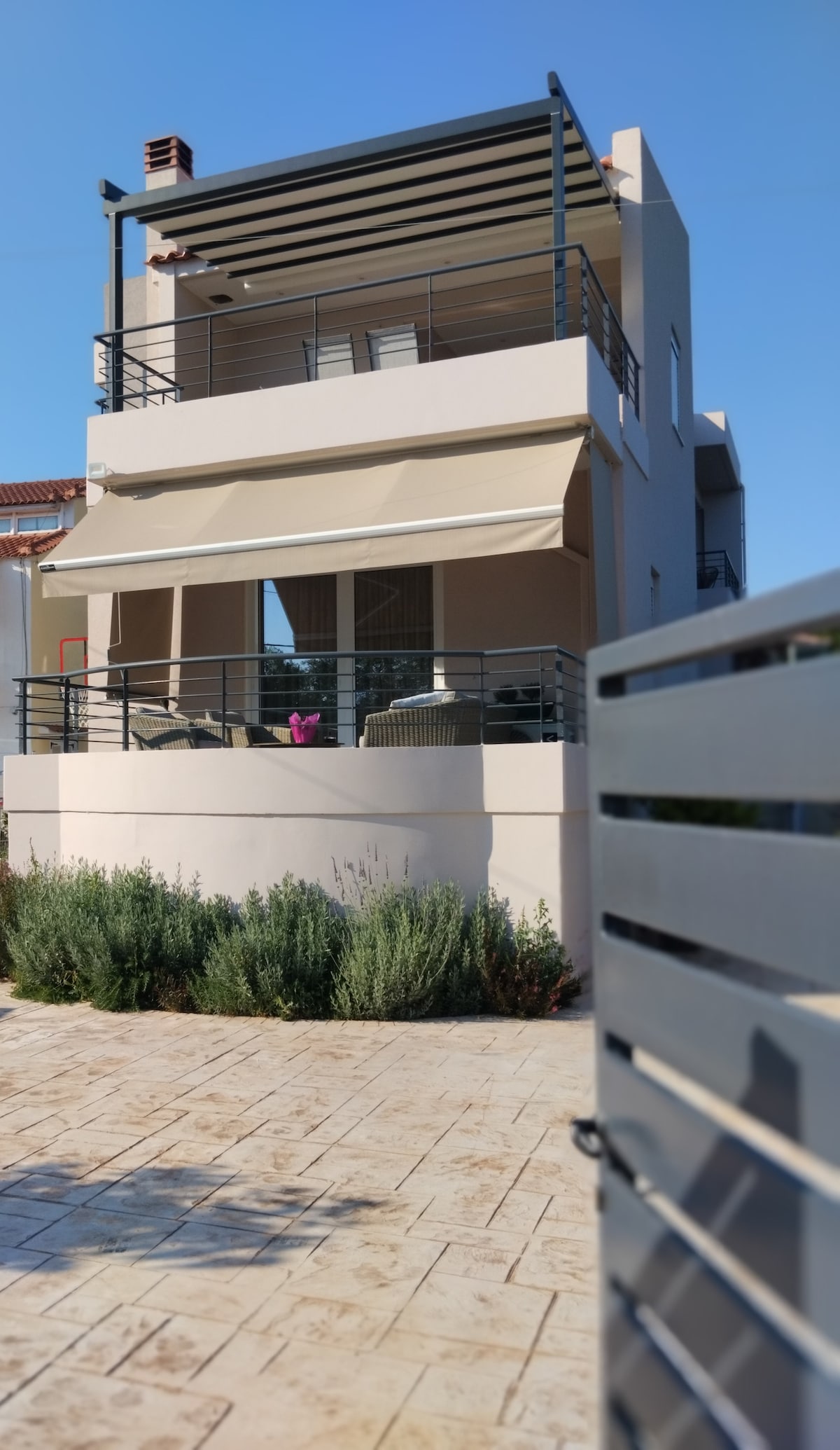 A newly built guesthouse is depicted, featuring a modern design with a spacious covered terrace and outdoor seating. Lush green shrubs are visible in the foreground, while the exterior is painted in neutral tones. The building is framed by clear blue skies.